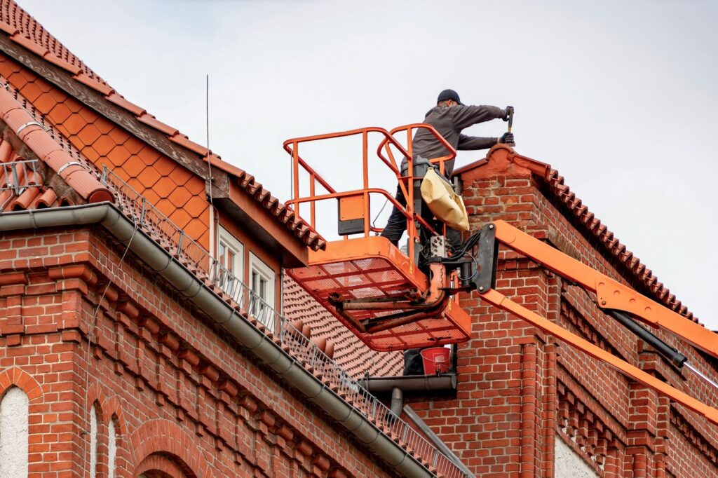 Ouvrier sur nacelle réparant le toit en tuiles d'un bâtiment en briques rouges.